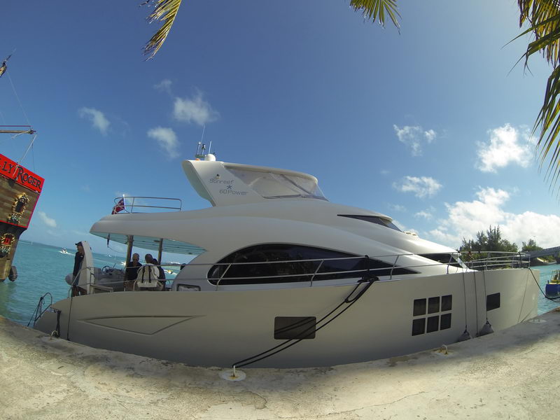 Side profile of a white Sunreef 60 Power catamaran moored along a stone quay framed by palm fronds, guests and crew visible on the aft deck, small boats and turquoise lagoon behind, deep blue sky.