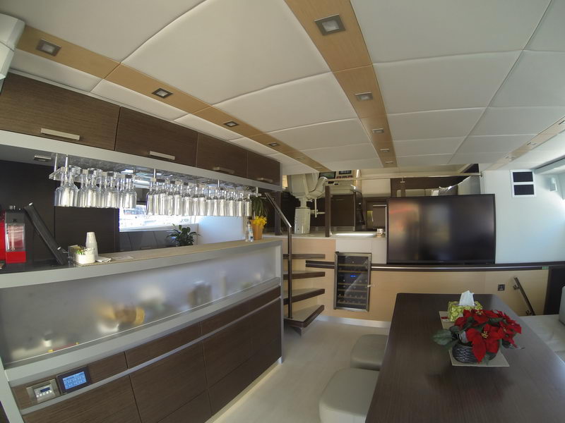 Catamaran salon interior with long illuminated bar, racks of hanging glasses, dark wood cabinetry, wine cooler under stairs, dark dining table with poinsettia centerpiece, padded white ceiling panels.