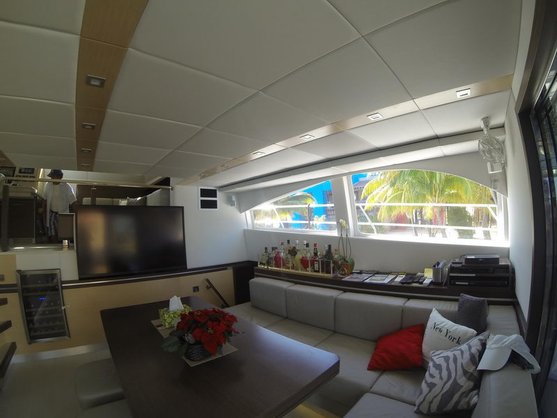 Catamaran salon with dark wood dining table and poinsettia arrangement, large TV on partition, gray sectional sofa with red and zebra cushions, bottle-lined shelf under wide windows showing palm trees.