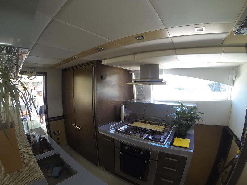 Catamaran galley with stainless four-burner gas cooktop, built-in oven, chimney extractor hood, dark wood cabinetry and fridge column, potted plants, window to side deck where crew stand outside.