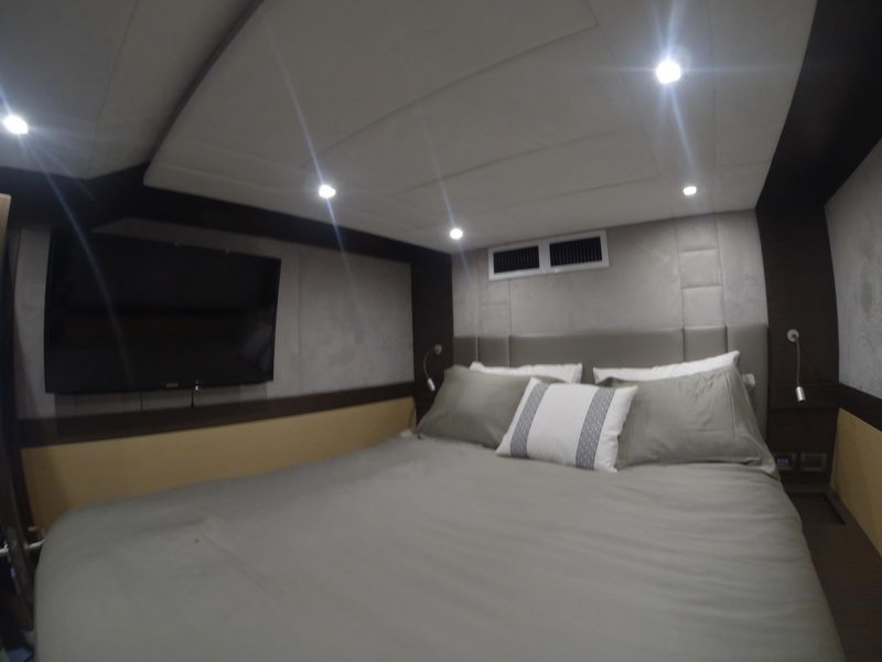 Guest cabin with double bed in gray linens and striped accent pillow, padded suede wall panels, wall-mounted TV, ceiling spotlights and air vent, warm yellow trim along the hull side.