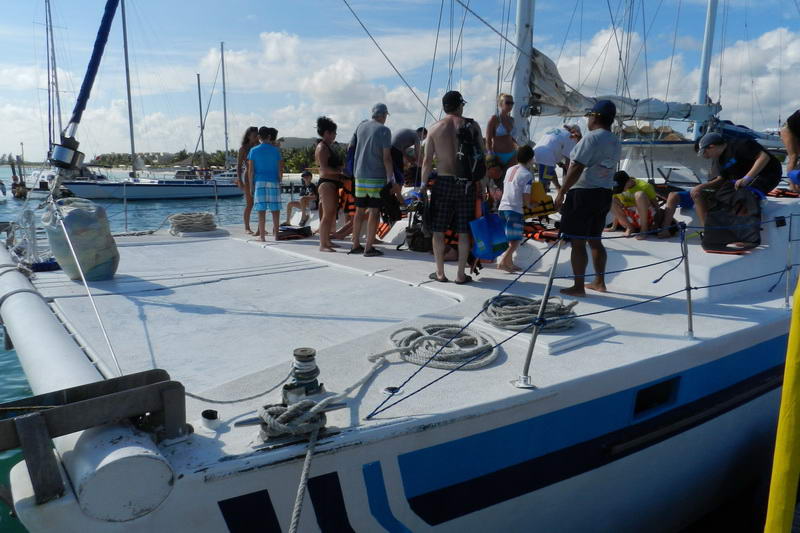 Invitados abordando la amplia cubierta de proa del catamarán Tahuna en el muelle de Maroma, con cabos enrollados sobre la red bajo un cielo azul