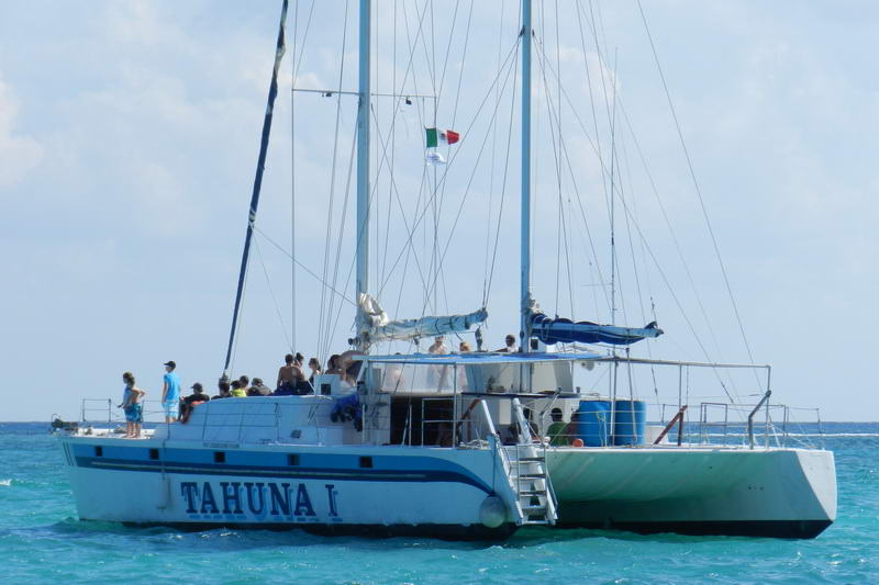 Catamarán de fiesta Tahuna I de casco azul fondeado en aguas turquesa del Caribe con invitados a bordo y la bandera mexicana, Maroma Playa del Carmen