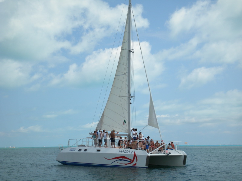 Side view of catamaran 'Samba 2' under full sail with two sails raised, passengers visible on deck, calm sea and partly cloudy sky.
